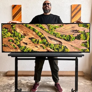 May include: A large rectangular wall art piece with a black frame. The artwork features preserved green and yellow moss, and natural wood elements. The wood has a light brown color and a unique grain pattern. The piece is displayed on a black table.
