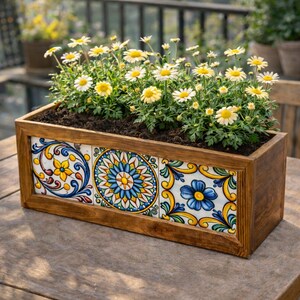 Puede incluir: Una jardinera rectangular de madera llena de tierra y margaritas en flor. La parte delantera de la jardinera presenta tres baldosas de cerámica decorativas con coloridos diseños florales en azul, amarillo y blanco. La jardinera está sobre una mesa de madera.