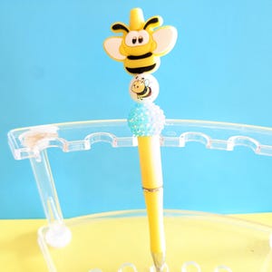 May include: A yellow pen with a bee-themed design. The pen features a large, cartoonish bee at the top, with a smaller bee on a white bead, and a blue beaded accent. The pen is displayed in a clear plastic holder against a yellow and blue background.