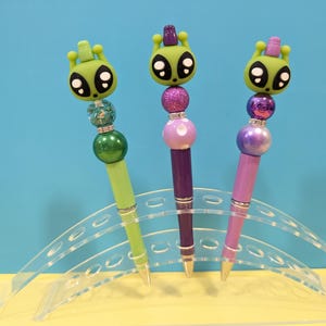 May include: Three decorative pens with alien-themed toppers. Each pen features a different color scheme: green, purple, and pink. The alien heads have large black eyes and antennae. The pens are displayed on a clear acrylic stand.