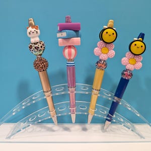 May include: Four decorative pens with unique toppers, including a cat, books, and bees. The pens have gold, pink, and blue barrels, and are displayed in a clear acrylic holder against a blue background. Each pen has a different decorative bead.