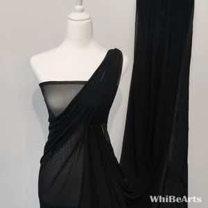 Op de afbeelding: Een zwarte jurk met één schouder, gedrapeerd op een witte mannequin. De jurk is gemaakt van een transparante, vloeiende stof, met een geplooid ontwerp op het lijfje. De jurk is vloerlang, met een bijpassend stoffen paneel aan de rechterkant. Het WhibeArts-logo is zichtbaar.