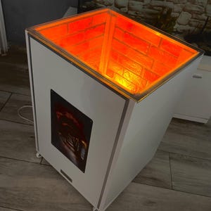 May include: White cube-shaped stand with a glass top. The top is illuminated with orange light and features a brick pattern. The stand has a black metal plate on the side with a picture of a dark, narrow hallway.