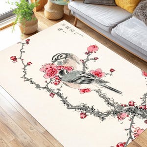 May include: Rectangular area rug with an Asian-inspired design. The rug features a cream-colored background with a detailed illustration of two birds perched on a flowering branch with pink blossoms. The rug is placed on a hardwood floor.