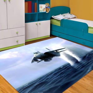 May include: A rectangular area rug featuring a detailed image of a jet fighter soaring over a turbulent ocean. The rug's design incorporates shades of blue and gray, creating a dynamic and realistic scene. The rug is suitable for a child's bedroom.