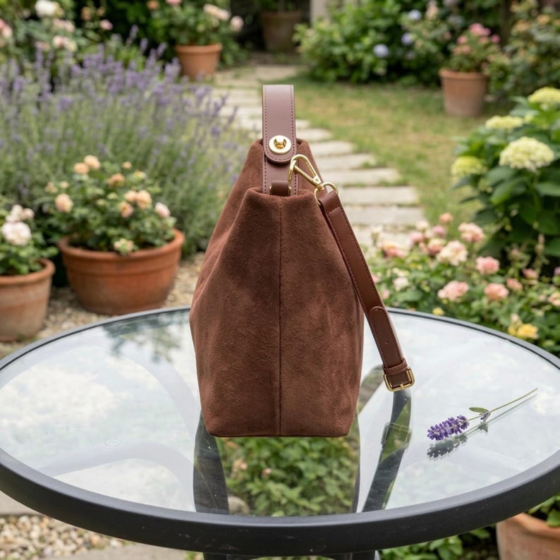 May include: A brown suede handbag with a top handle and a detachable shoulder strap. The bag has gold-colored hardware and is sitting on a glass table. The background features a garden with flowers and plants.