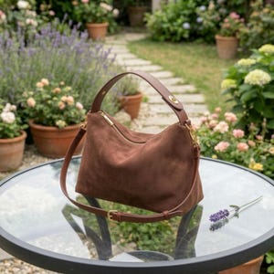 Puede incluir: Un bolso de hombro de ante marrón con asa superior y correa desmontable. El bolso tiene cremallera y herrajes dorados. El bolso está sobre una mesa de cristal con una ramita de lavanda.