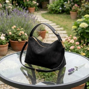 May include: Black suede shoulder bag with a gold zipper and hardware. The bag has a top handle and a detachable shoulder strap. The bag is sitting on a glass table with a garden in the background.