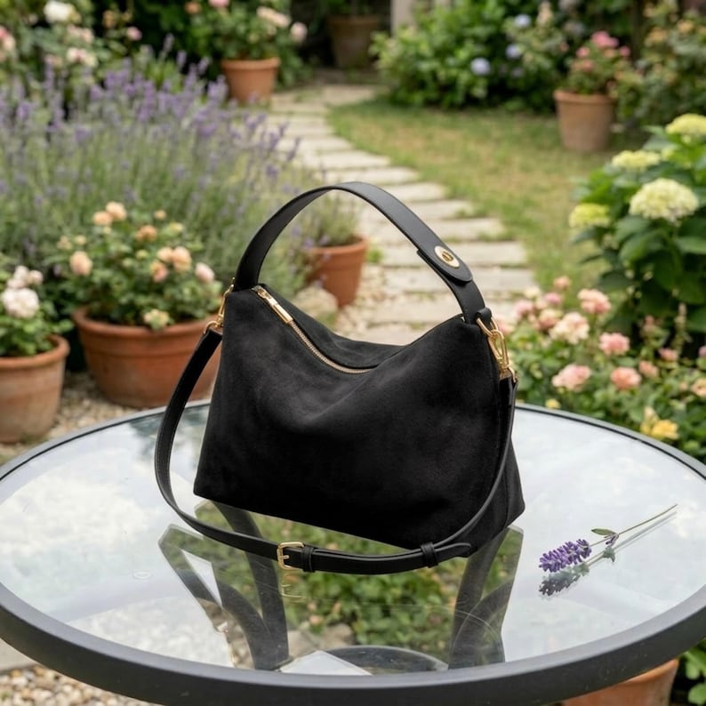 May include: Black suede shoulder bag with a gold zipper and hardware. The bag has a top handle and a detachable shoulder strap. The bag is sitting on a glass table with a garden in the background.