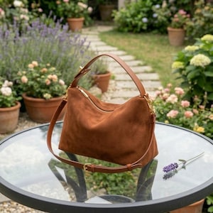 May include: A brown suede shoulder bag with a gold zipper and brown leather straps sits on a glass table. The bag has a top handle and a longer adjustable strap. A sprig of lavender rests on the table.