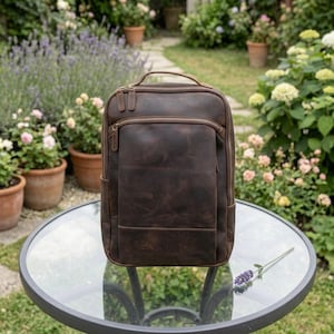 Puede incluir: Una mochila de cuero marrón oscuro con múltiples compartimentos con cremallera y un asa superior. La bolsa tiene un acabado envejecido y una forma rectangular. El fondo muestra un jardín con plantas en macetas y flores.