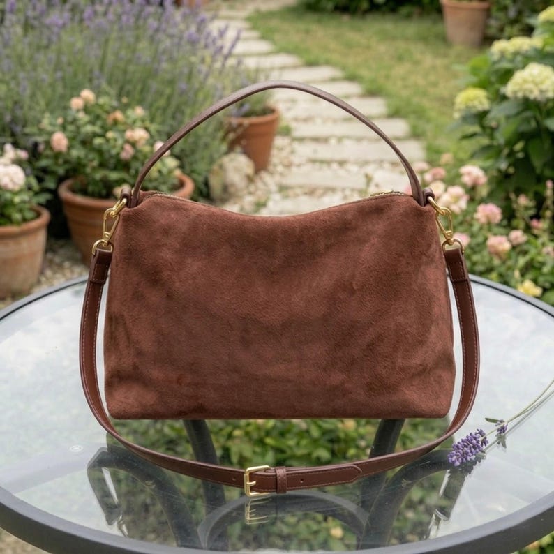 May include: A brown suede handbag with a top handle and a detachable shoulder strap. The bag has gold-colored hardware and a rectangular shape. The bag is sitting on a glass table in a garden setting.