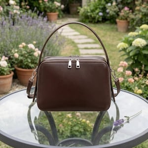 May include: A dark brown rectangular handbag with silver zips and fittings. It features a top handle and a detachable shoulder strap. The bag is on a glass table, with a garden visible in the background.