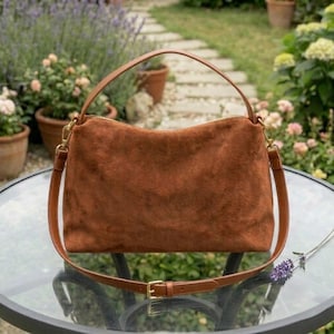 May include: A brown suede handbag with a top handle and a detachable shoulder strap. The bag has a rectangular shape with a zipper closure and gold-colored hardware. The strap is adjustable with a gold-colored buckle.