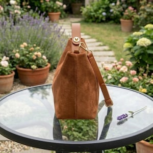 May include: A brown suede handbag with a leather handle and shoulder strap. The bag has a gold-colored clasp and is sitting on a glass table. The background features a garden with flowers and potted plants.