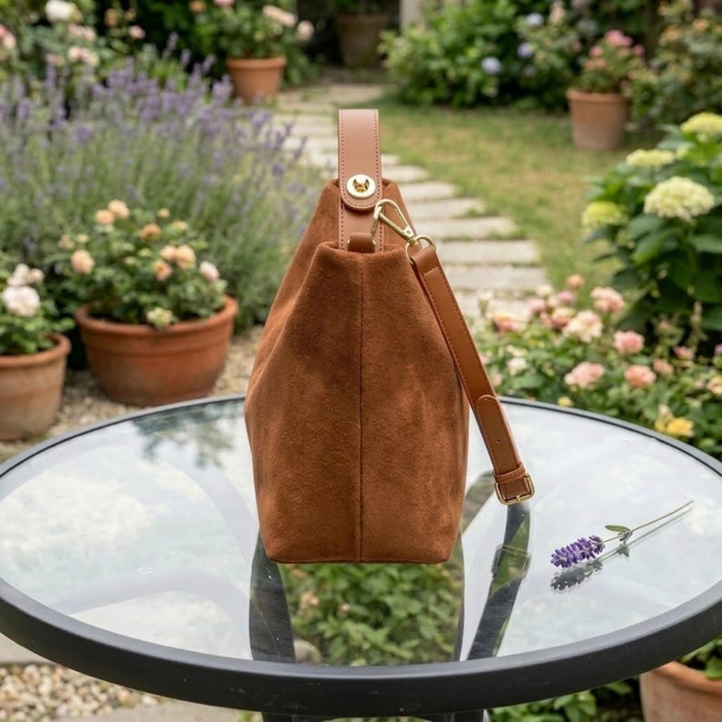 May include: A brown suede handbag with a leather handle and shoulder strap. The bag has a gold-colored clasp and is sitting on a glass table. The background features a garden with flowers and potted plants.