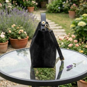 May include: A black suede handbag with a structured design. It features a top handle and a detachable shoulder strap. The bag has gold-tone hardware and is set on a glass-topped table in a garden setting.