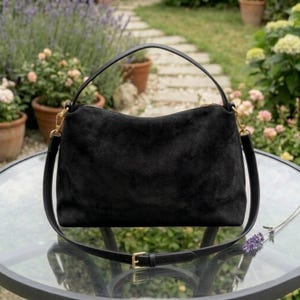 May include: A black suede handbag with a top handle and a detachable shoulder strap. The bag has gold-colored hardware and a zipper closure. The bag is sitting on a glass table in a garden setting.
