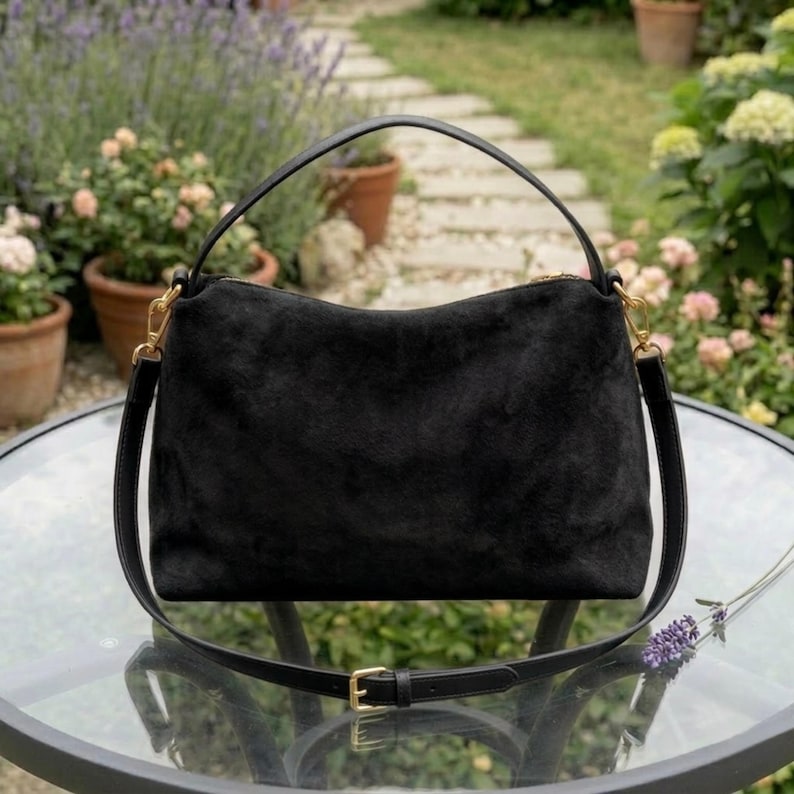 May include: A black suede handbag with a top handle and a detachable shoulder strap. The bag has gold-colored hardware and a zipper closure. The bag is sitting on a glass table in a garden setting.