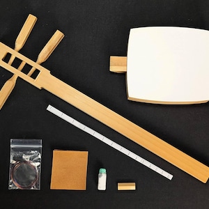 Op de afbeelding: Een gedemonteerde shamisen, een traditioneel Japans driestrengs instrument, met een lichte houten afwerking. De kit bevat een witte klankkast, een liniaal, snaren en andere componenten, alles tegen een zwarte achtergrond.