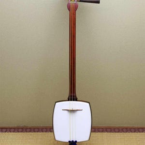 Puede incluir: Un instrumento de cuerda japonés tradicional, el shamisen, que presenta un mástil de madera largo y delgado, un cuerpo cuadrado con una caja de resonancia cubierta de piel blanca, tres cuerdas amarillas y clavijas de madera oscura.