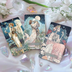 May include: Three tarot cards with skeleton figures in a gothic style. The cards, labelled "Temperance," "The Lovers," and "The Empress," show scenes with floral and celestial elements. Crystals and white flowers surround the cards.