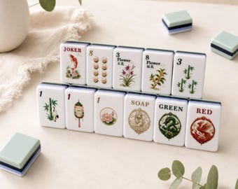 May include: A set of rectangular mahjong tiles with white faces and dark blue backs. The tiles feature various colourful illustrations including a koi fish, bamboo, flowers, a lantern, a lotus, a cat, a dragon, and a phoenix, along with text labels.