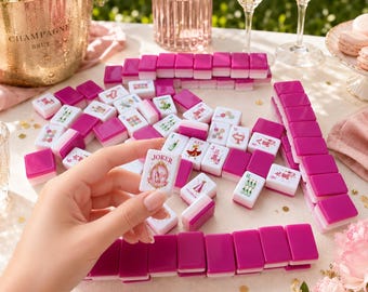 May include: A set of Mahjong tiles featuring a vibrant fuchsia pink and white color scheme. The tiles are arranged in rows and scattered across a surface, with one tile held up displaying a Joker design with a champagne bottle illustration.