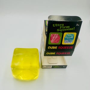 May include: A translucent yellow cube-shaped sensory toy next to its black and clear packaging. The box features the text Ultra Firm Squeeze! and CUBE SQUEEZE, along with images of pink and blue marbled cubes. The packaging indicates a 3+ age rating.