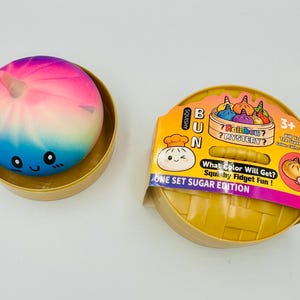 Puede incluir: Un juguete squishy redondo de colores arcoíris con cara sonriente y un pequeño cuerno, dentro de un recipiente dorado. Al lado hay un recipiente dorado tipo cesta con la etiqueta Squishy Bun Rainbow Mystery One Set Sugar Edition.