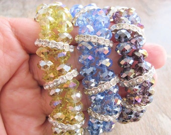 May include: Three stretch bracelets featuring faceted crystal beads in yellow, light blue, and dark purple. Each bracelet is accented with silver-toned spacers embedded with small clear rhinestones, arranged in a repeating pattern along the strand.