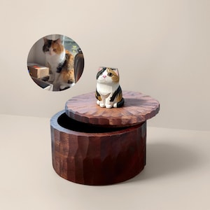 Pode incluir: Uma caixa de madeira artesanal com exterior entalhado e tampa. Uma pequena figura de gato de madeira pintada está em cima da tampa. O gato é branco, preto e castanho. A caixa é castanha escura e tem formato redondo.