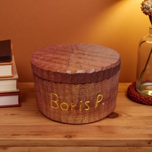 May include: A round wooden box with a lid, inscribed with "Boris P." in gold lettering. The box is crafted from light brown wood with a darker brown rim on the lid. It rests on a wooden surface.