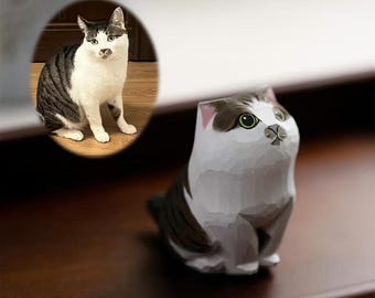 May include: A hand-carved wooden cat figurine in a sitting position, painted white and dark gray. The cat has green eyes and a small black nose. An oval image of a real cat is in the background.