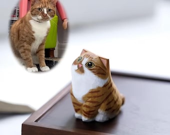 Puede incluir: Figura de gato de madera tallada a mano con un patrón atigrado marrón y blanco. El gato tiene ojos grandes y expresivos y está sentado sobre una superficie de madera oscura. Una foto de un gato real está en el fondo. Un artículo decorativo encantador.