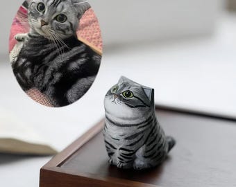 May include: A wooden figurine of a gray and black striped cat with green eyes. The cat is sitting upright with its head tilted upwards. An oval photo of a similar cat is in the background.