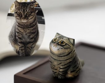 May include: A wooden cat figurine with a tabby pattern, featuring black stripes on a gray base. The cat has large yellow eyes and is sitting on a dark brown wooden surface. A real cat is reflected in the background.