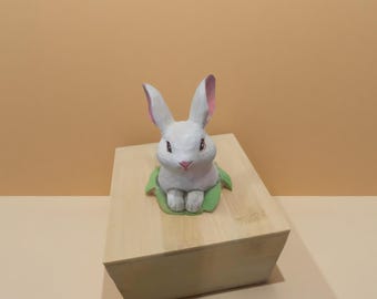 Puede incluir: Una figura de conejo blanco con el interior de las orejas rosa y una nariz rosa, sentado sobre una hoja verde, encima de una caja de madera marrón claro. El conejo está pintado a mano y tiene un aspecto caprichoso y caricaturesco.