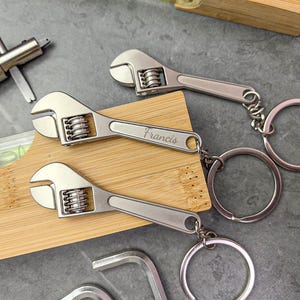 Może przedstawiać: Srebrne breloczki do kluczy w kształcie klucza nastawnego z kółkami. Na jednym breloczku wygrawerowano imię "Francis". Breloczki leżą na drewnianym bloku i szarej powierzchni. Na zdjęciu widoczne są również inne narzędzia.