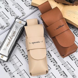 Puede incluir: Una armónica plateada con el nombre "maddison" grabado, junto a dos estuches de cuero. Un estuche es beige y el otro marrón, ambos con nombres grabados. Los artículos están sobre una partitura musical.