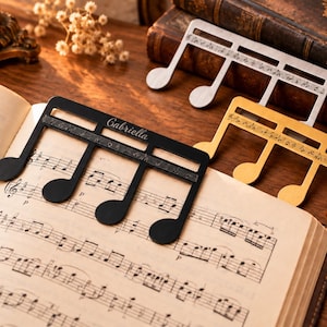 May include: Three metal music note bookmarks in black, gold, and silver finishes. Each bookmark features a horizontal staff with engraved musical notes. The black bookmark is engraved with the name Gabriella and rests on an open music book.
