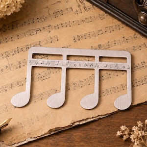 Puede incluir: Un adorno de metal plateado con forma de semicorchea, que presenta una l&iacute;nea de pentagrama horizontal grabada en el centro con varias notas musicales y s&iacute;mbolos, incluyendo una clave de sol y notaciones r&iacute;tmicas grabadas en la superficie.