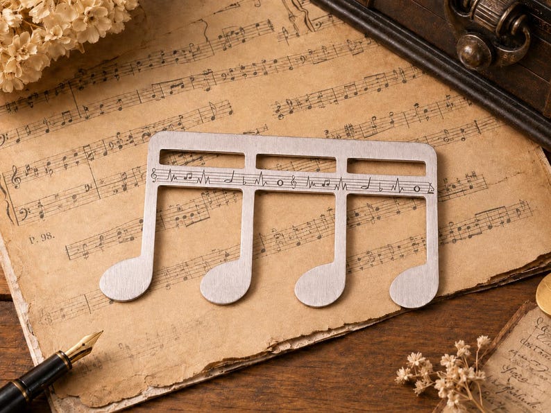 Puede incluir: Un adorno de metal plateado con forma de semicorchea, que presenta una l&iacute;nea de pentagrama horizontal grabada en el centro con varias notas musicales y s&iacute;mbolos, incluyendo una clave de sol y notaciones r&iacute;tmicas grabadas en la superficie.
