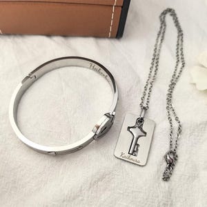 May include: A silver-colored bracelet and necklace set. The bracelet has the word "Habeeba" engraved on it. The necklace features a key-shaped pendant with the word "Kaikoura" engraved on it. A white flower is in the background.