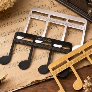 Puede incluir: Tres sujetap&aacute;ginas de metal con forma de nota musical en acabados plateado, negro y dorado. El sujetap&aacute;ginas negro tiene grabado el nombre Gabriella en letra cursiva. Cada pieza incluye una barra horizontal con pentagramas musicales grabados.
