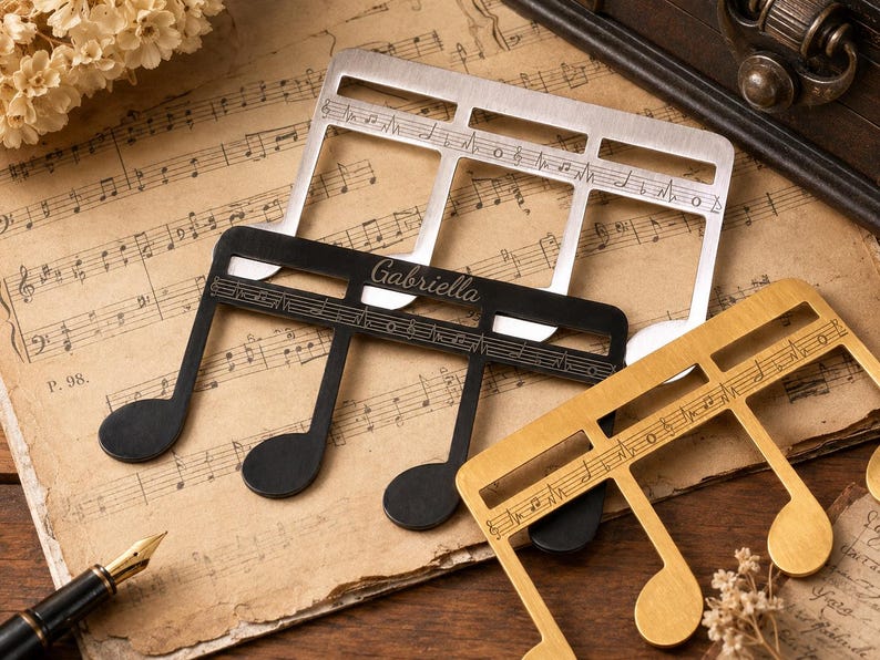 Puede incluir: Tres sujetap&aacute;ginas de metal con forma de nota musical en acabados plateado, negro y dorado. El sujetap&aacute;ginas negro tiene grabado el nombre Gabriella en letra cursiva. Cada pieza incluye una barra horizontal con pentagramas musicales grabados.