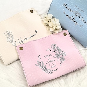 May include: Four personalized catch-all trays in cream, pink, and blue. Each tray features a unique design and text, including floral illustrations and names with dates. The trays are fastened with brass-colored snaps.