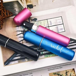 Puede incluir: Varios atomizadores de perfume en negro, azul y rosa. El atomizador azul está inscrito con el nombre "rachel" en escritura cursiva. Cada atomizador tiene una banda plateada y una tapa a juego. Los atomizadores se muestran sobre una superficie impresa.