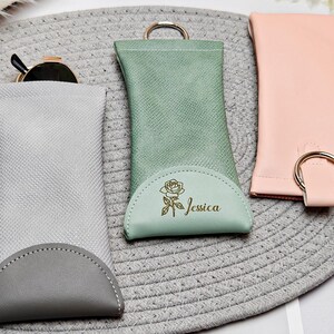 May include: Three eyeglass cases in a row. The first is light grey with a darker grey bottom, the second is mint green with a rose and the name "Jessica", and the third is pink. All have a ring.