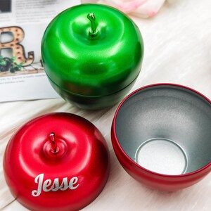 May include: Three apple-shaped containers, one green and two red. The open red container reveals a silver interior. The other red container has the name "Jesse". The green container is closed.